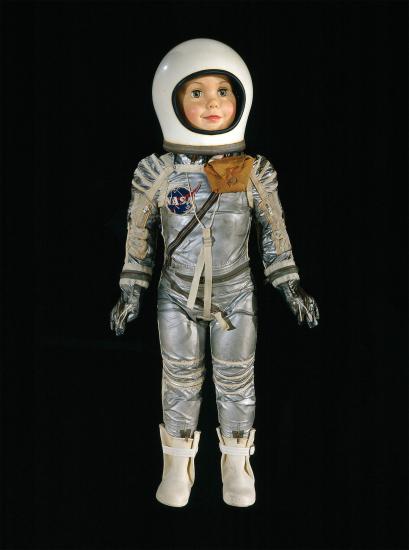 A 3-foot-tall freckle-faced plastic doll is dressed as an astronaut. The doll is wearing a white plastic helmet, a silver spacesuit, and white rubber boots.