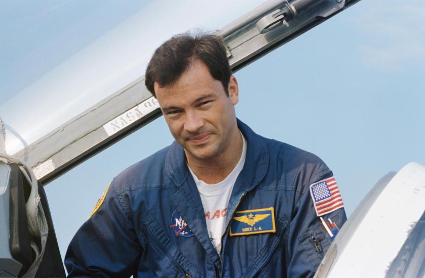 An individual in a blue flight suit with NASA and American flag patches, standing next to part of an aircraft with the sky in the background. 