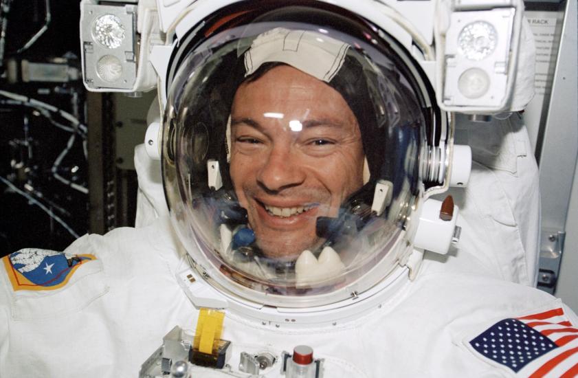 An astronaut wearing a spacesuit and smiling at the camera. The suit features patches of the American flag and another emblem with a star. The background shows equipment and parts of a spacecraft interior, suggesting this image was taken during a space mission.