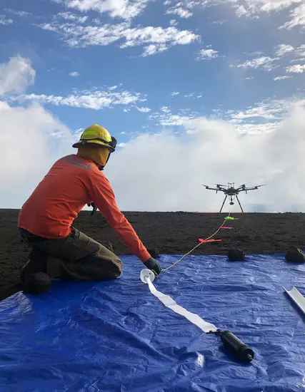 A person wearing a yellow safety helmet and a long-sleeve orange t-shirt crouches on the ground, preparing to launch a drone.