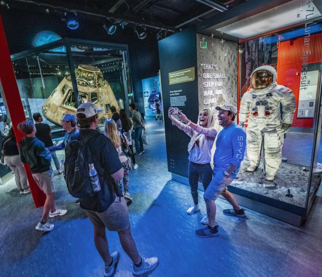 A group of people in the Destination Moon gallery. Two people take a selfie with Neil Armstrong's spacesuit, which is in a case.