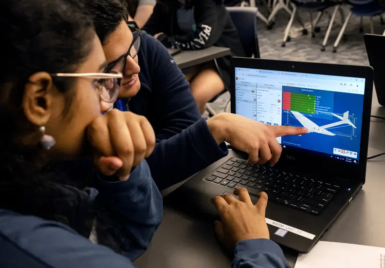 A man with glasses points to a schematic of an airplane on a laptop. A young woman sits behind the laptop. 