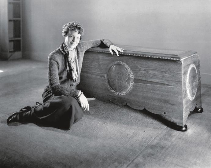 A slender white woman in her late 30s sits on the floor inside a house in this black and white photo. She has one arm draped over a large, hand-carved wood chest. The woman has short curly brown hair. She is wearing a long-sleeve sweater and a long dark skirt. A silk scarf is tied around her neck.