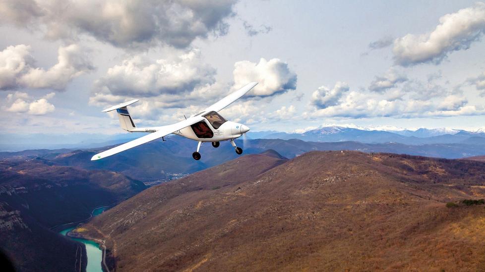 A small, white airplane flies low over a river and brown desert hills. Large white clouds are above the airplane. The airplane is a high-wing monoplane with a single propeller-driven engine and tricycle landing gear.