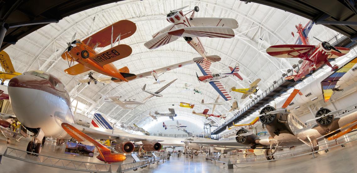 Dozens of various aircraft of all type hang suspended from the ceiling in a large hangar. More aircraft dot the ground.