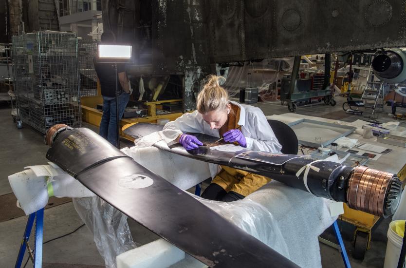 A woman with blonde hair, leans down in intense focus on the propeller of an airplane. The rest of a the conversation hangar fades out around her, you can feel her focus. 