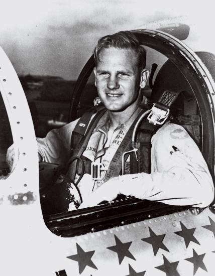 A white man in his 30s sits in the cockpit of a military airplane in this black-and-white image. The plexiglass canopy covering the cockpit is slid open, showing how the man is strapped in tightly.