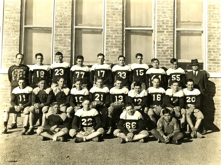 A vintage group photograph of a high school football team with players in jerseys and a coach, posing in front of a brick building.