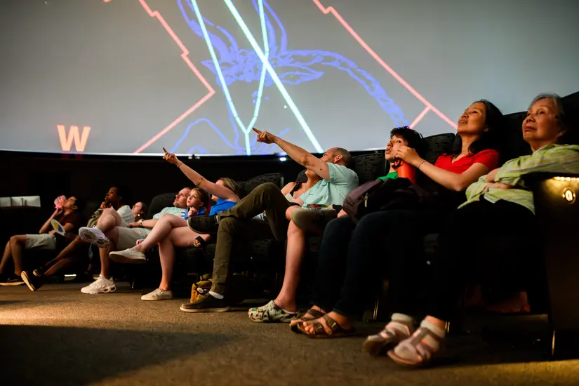 People sitting in seats in a planetarium. Constellations can be seen on the domed screen.