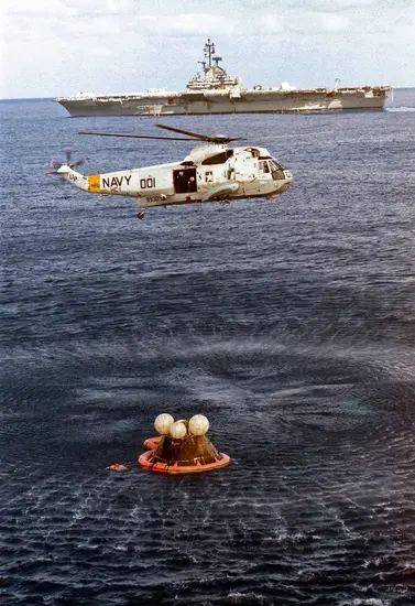 A Navy helicopter hovers over the ocean near a life raft with two individuals, with a large naval ship visible in the background.