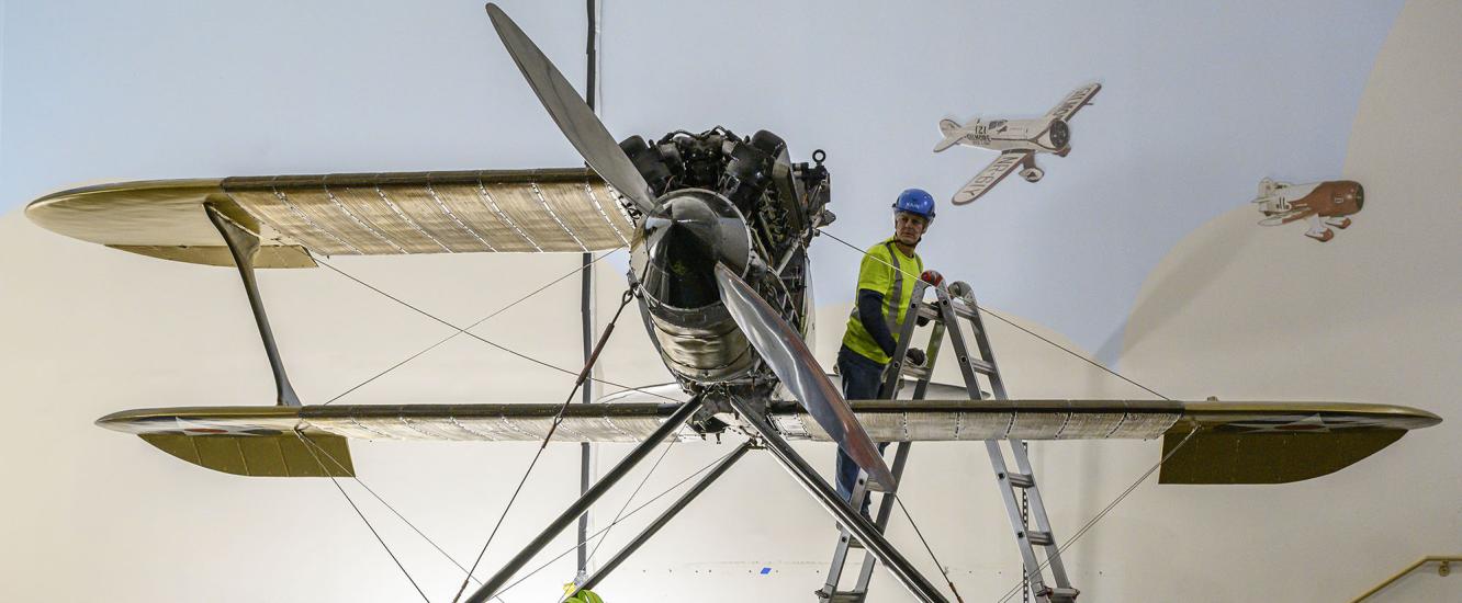 A worker stands on a ladder looking at the Curtiss R3C-2 Racer, which has one wing removed