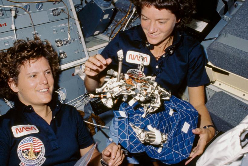 Sally Ride and Kathryn Sullivan, both of whom are white female astronauts, display an object in space.