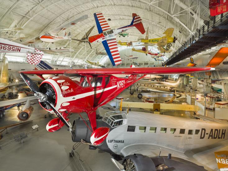 Red sporting monoplane with white trim and clipped wings on display in aviation hangar