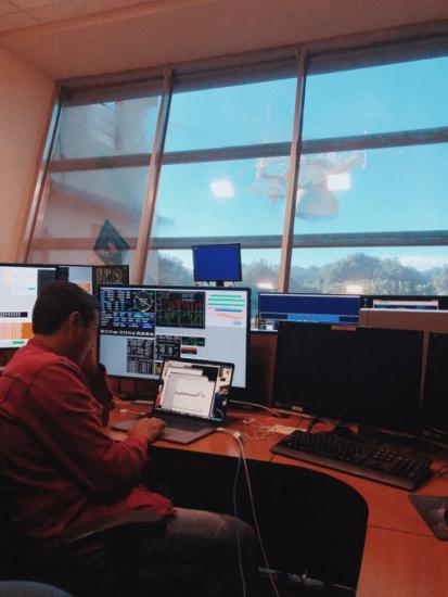 Young man sitting at a desk with computers. Telescope of the Arecibo Observatory can be seen out the window.