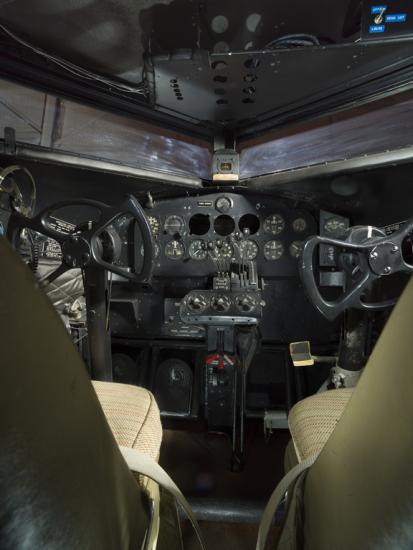 View of the cockpit on the plane. The image was taken behind the seats of the cockpit. The image highlights the instrument panel of the plane. 