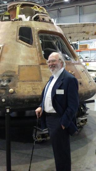 Board member Bruce R. McCaw, an older white male with a beard, poses informally in front of the Command Module Columbia