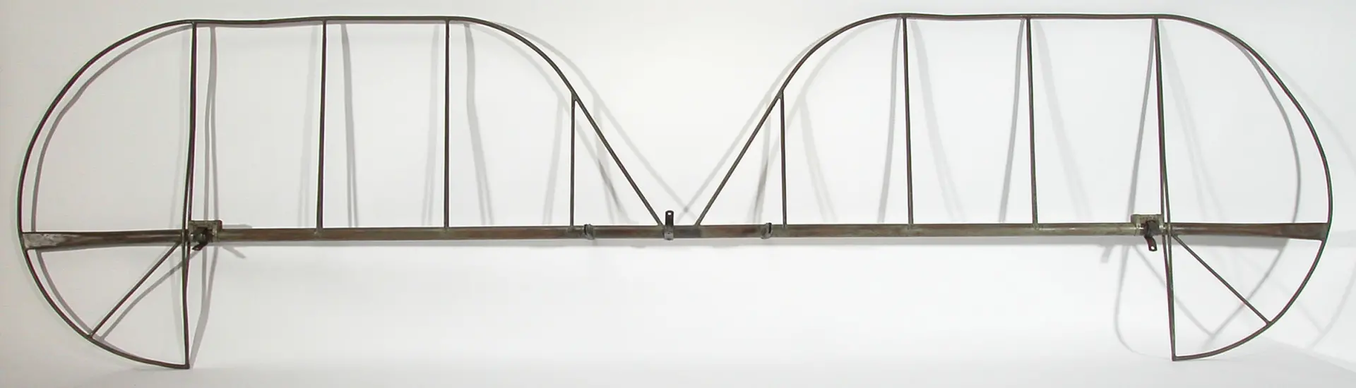 A skeletal metal frame shaped like an airplane tail section, featuring two symmetrical, curved vertical stabilizers connected by a central horizontal bar. The structure is composed of thin metal rods arranged in triangular and rectangular patterns, with no outer covering or additional parts attached.