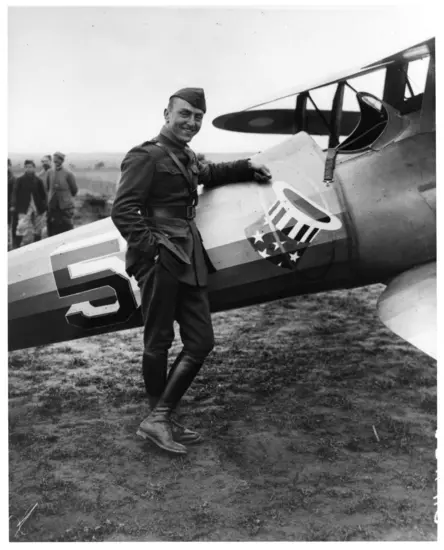 A white man in military uniform smiles while standing in a relaxed pose with his arm around his airplane. A star-spangled top hat is painted on the body of the aircraft. 