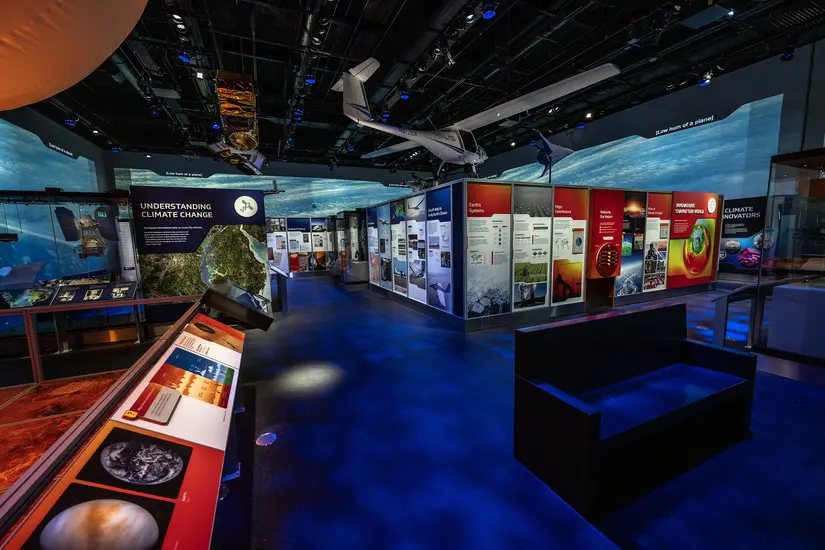 A photograph taken in the far back corner. Moving from left to right, in the foreground is a weather balloon illuminated orange, mirroring the environment on Venus. In the center of the gallery are panels with text and images. One panel has the heading "Understanding Climate Change." Artifacts hang above the panels, including a satellite and small plane. In the front right corner is a bench. 