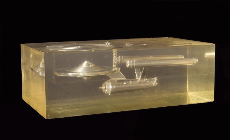 A model of the saucer shaped starship Enterprise made of a silver-tone metal (steel) in a lucite block with a partial beaded chain imbedded in the interior toward the top of the block. The rest of the chain is above and outside block, missing.