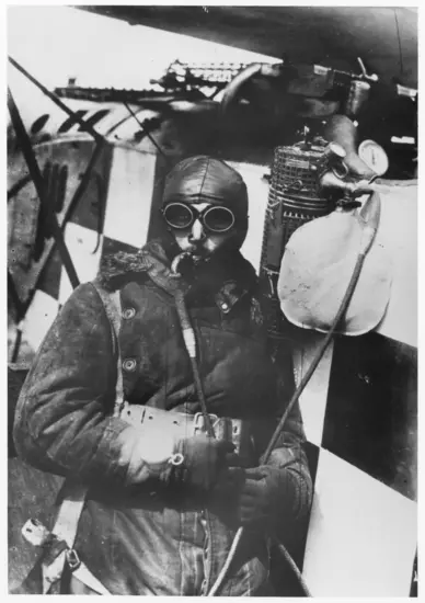 A man in heavy flying clothes with a leather helmet and goggles poses in front in an aircraft with ah checkered design on its surface. He has a tube in his mouth that is attached to a canister that is hanging off of his plane. 