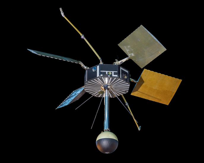 Short cylindrical satellite with a number of square solar panels extending off it and a sphere extended on a pole off the bottom of it.