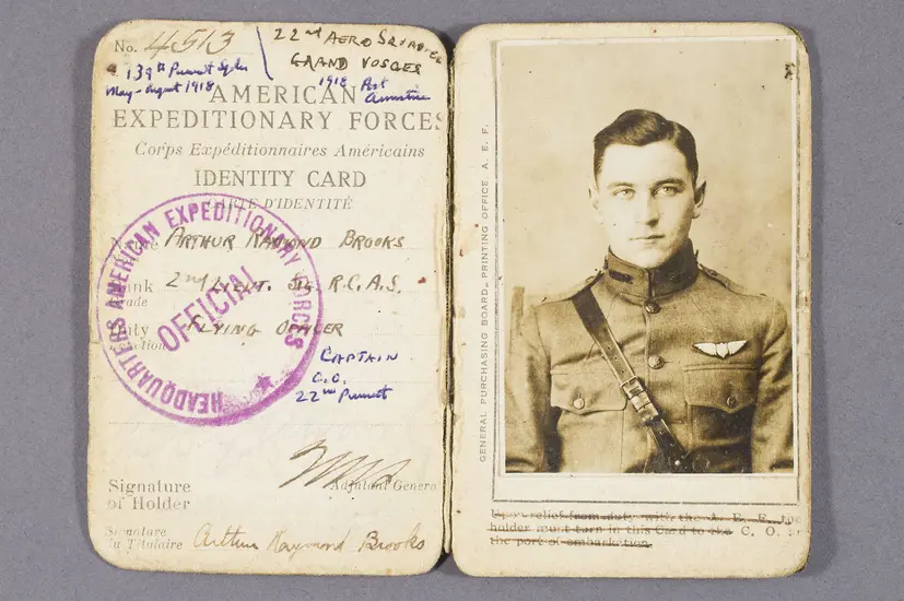 A manilla paper identification card, open to reveal a striking photograph of a young white man in a World War I military uniform. The left side of the page includes printed text reading "American Expeditionary Forces," handwriting including the name "Arthur Raymond Brooks" and "Flying Officer." Overtop the text is a purple circular stamp labeled "Official."