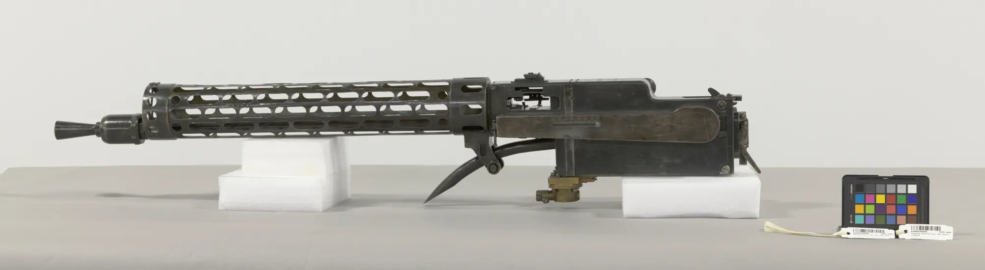 Black metal gun with a circular perforated barrel jacket, and muzzle booster on barrel. A synchronizer attachment is on the bottom of the gun.