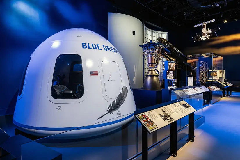White capsule with blue trim at base, painted feather logo, "Blue Origin" lettering in blue, and a U.S. flag all near the hatch sits in the foreground of the image, followed by a variety of technology--much used in commercial spaceflight. 