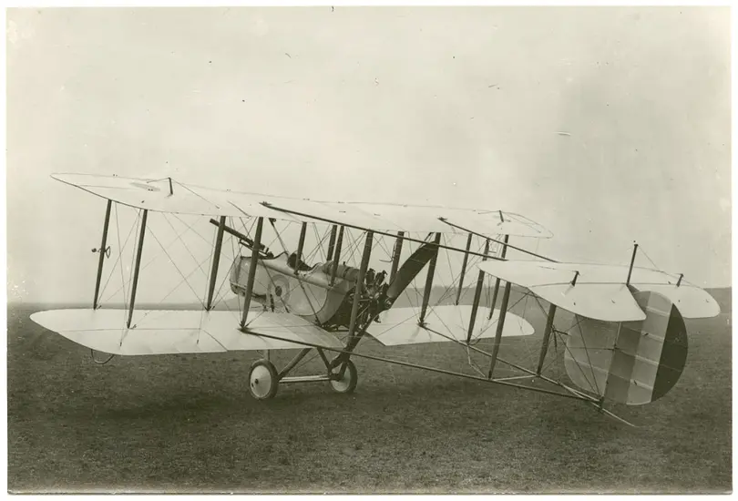 A black and white photograph of a biplane with a long gun mounted to the front. 