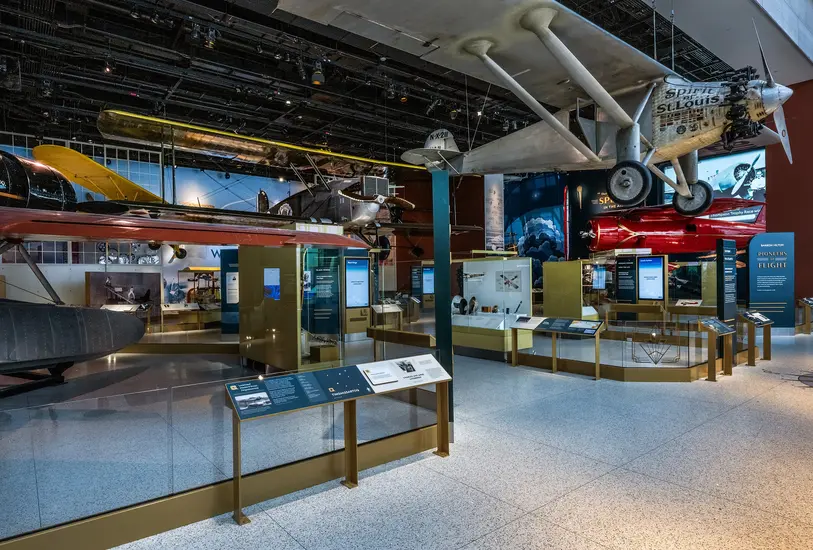 A Museum gallery full of 1920s and 1930s airplanes. Hanging from the roof at the entrance is a silver airplane with a propeller. Text on the side of the plane read "Spirit of St. Louis." Behind it, more aircraft is evident, including a biplane and a small red airplane. There are also metal cases to hold objects. 