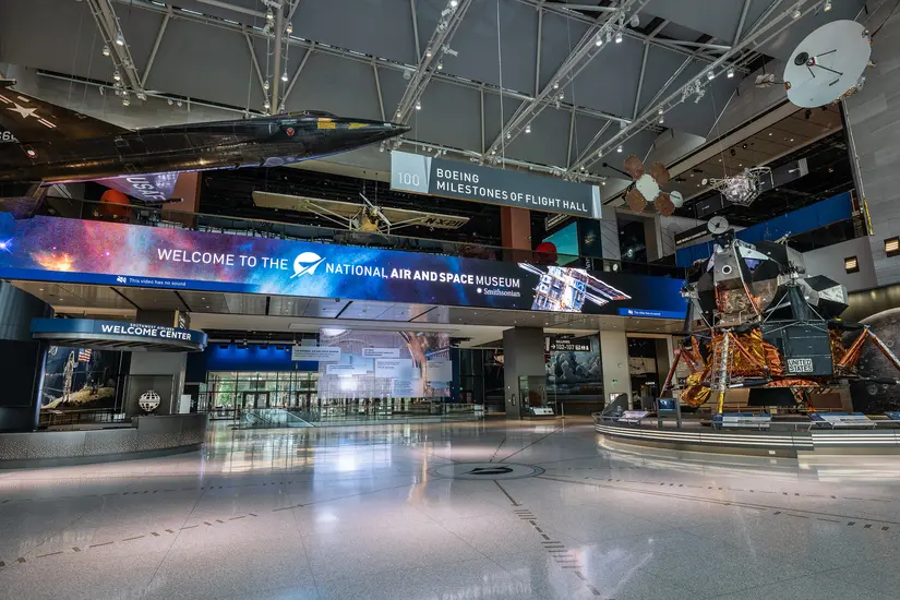 A museum gallery showing highlights from the Museum's collection, including an experimental black plane, a lunar module, satellites, and an electronic sign welcoming visitors the museum. 