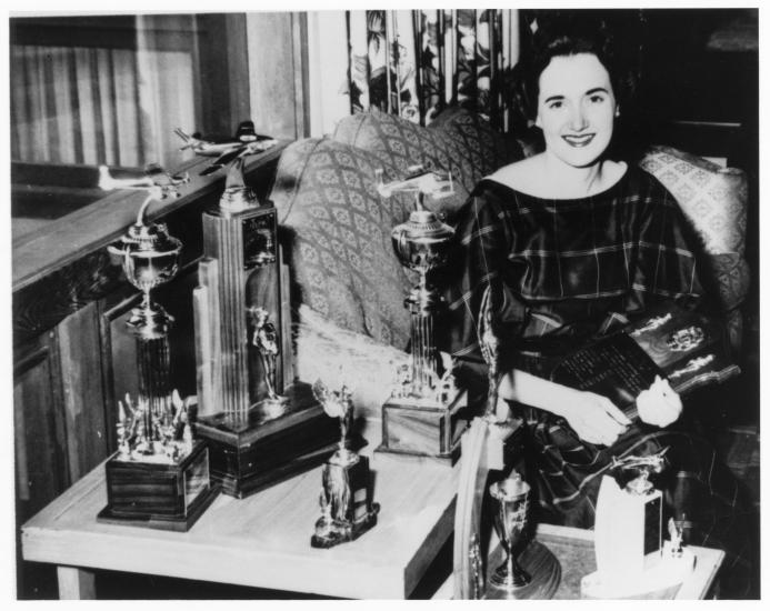 Black and white image of a woman sitting by a desk with various trophies.