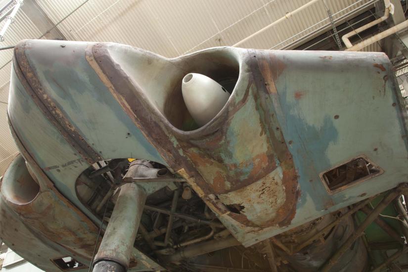 A deteriorated bottom section of a World War II era aircraft shows issues with paint loss, paint discoloration, thinning of metal, and coatings of preservatives visible across the aircraft.