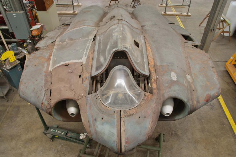 Top view of a gray, World War II era aircraft. On the left side, fairings (material covering gaps on an aircraft) on the aircraft are more rusted in comparison to the right side.