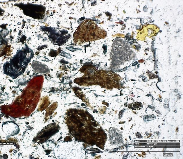 A microscopic view of a thin section of adhesive sampled from a World War II era aircraft, with various particles of various colours intertwined with the adhesive.