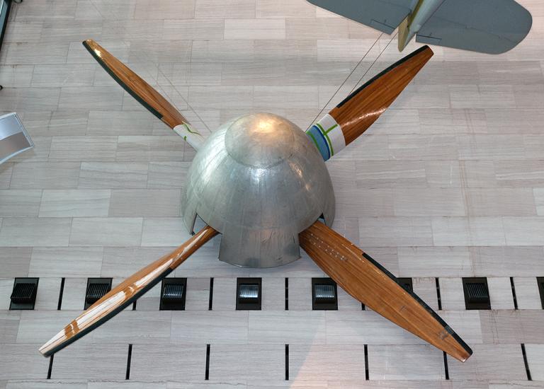 Large, four-blade wooden fan used for a wind tunnel that is hanging in the museum. The center is a silver-colored metal base.