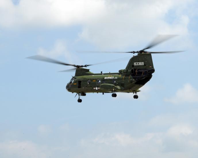 Side view of green military helicopter in flight. The helicopter has two separate propellors, both of which have three blades.