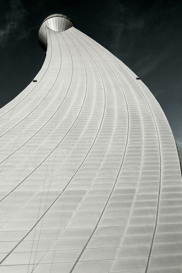 Partial view of the air traffic control tower at Abu Dhabi International Airport. The base has a curved, white tile pattern with the top leading to a metal control tower base.