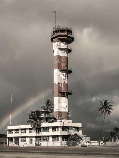 Partial view of an air traffic control tower at Ford Island Airport in Hawaii. The base features an alternating white and brown-colored concrete with three open balconies. Above the base, a single story exists for monitoring aircrafts and a balcony is found throughout the top of the tower.