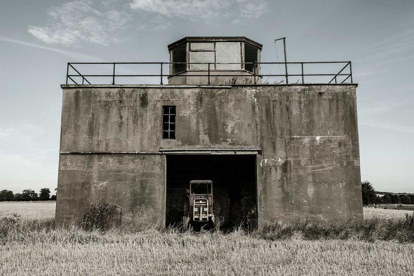 Partial view of the air traffic control tower at East Fortune Field in Scotland. The tower is three stories and features a concrete base with an opening in the center of the first floor. The third floor features at least four different observation angles.