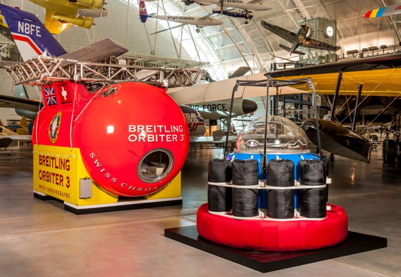 A large red and yellow balloon gondola with a rectangular shape is next to a red and black gondula with a smaller, red circular base and black supports on top of the base. They are both placed in the Museum's Steven F. Udvar-Hazy Center.