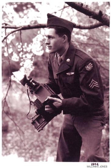 Sgt William E. "Bill" Jones, a white male U.S. Army photographer, crouches as he holds a camera in a forest.