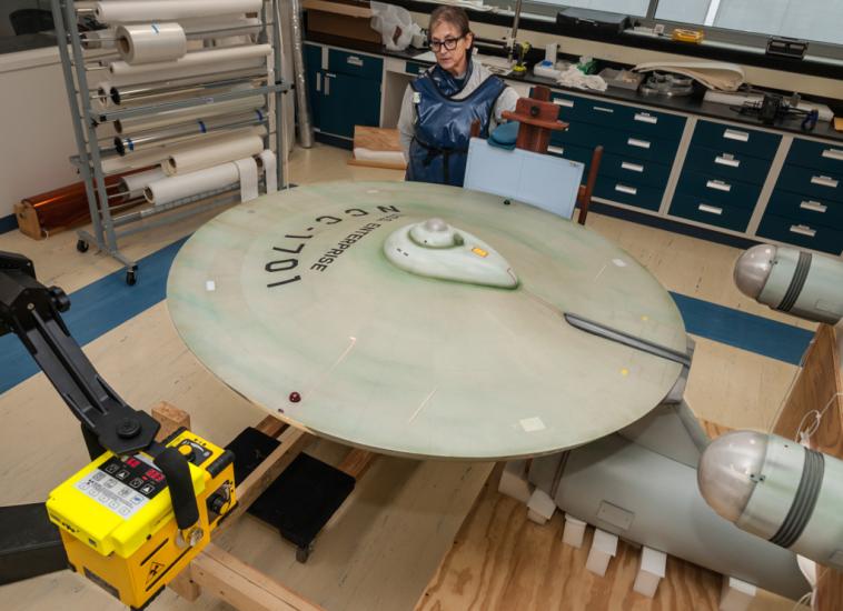 Top view of the starship Enterprise, a gray-colored, dish-shaped starship studio model, with a Smithsonian staff member standing near the studio model. The studio model is undergoing an X-ray scan.