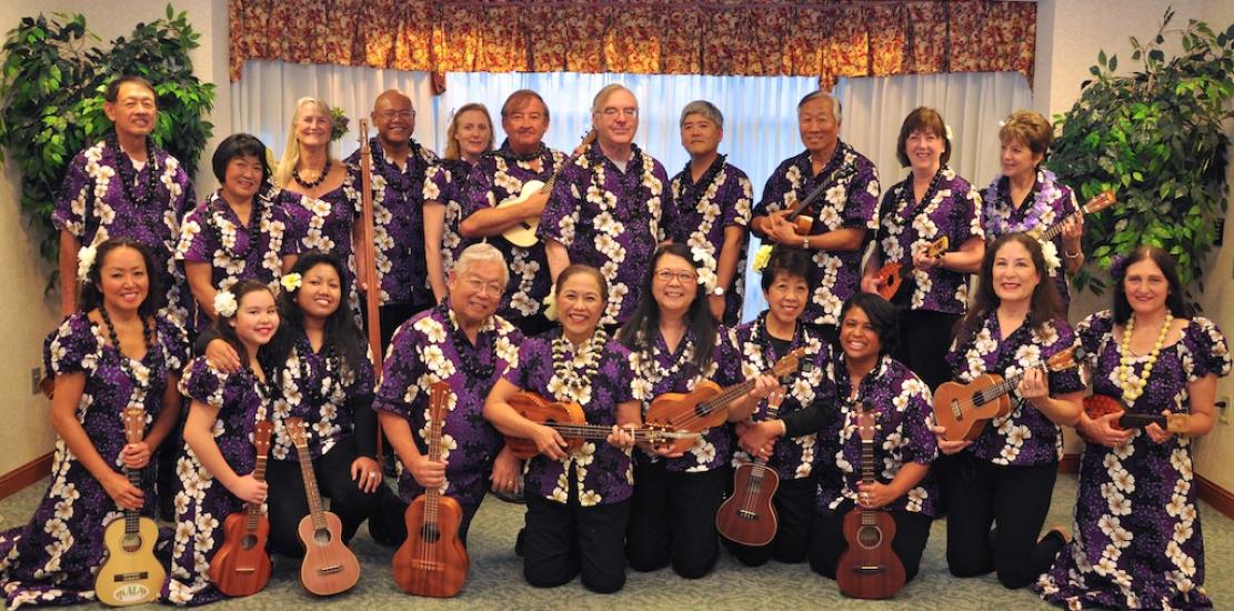 A large ensemble of ukulele players pose together with their ukuleles.