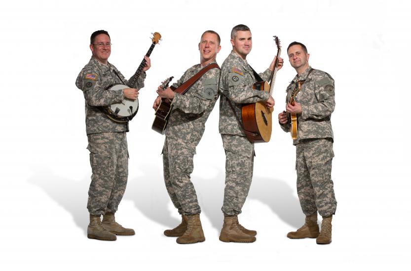 A group of four men in Army uniform pose informally with their instruments. The person on the far left is playing a banjo while everyone else seems to be playing guitar.