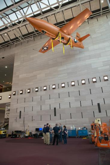 Several Museum employees stand under an orange monoplane hanging on the ceiling of the Museum's Boeing Milestones of Flight Hall. Nearby, an orange machine used to lift employees toward the aircraft is stationary.