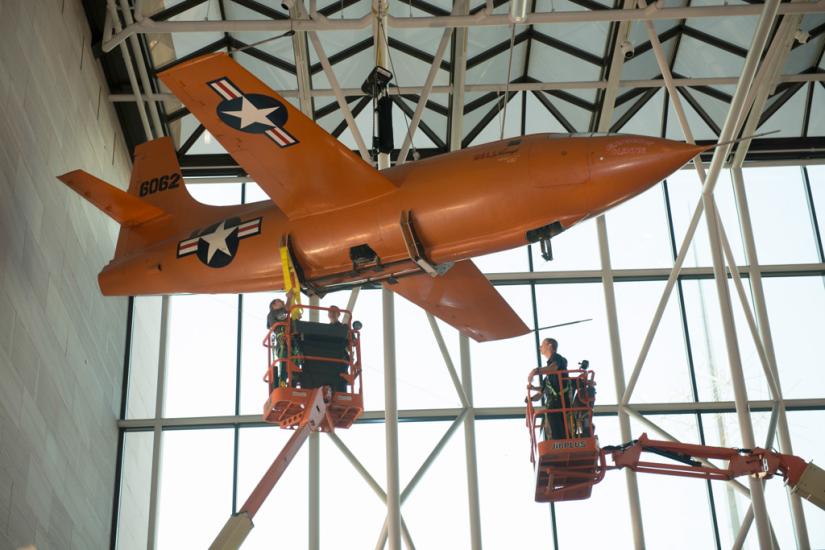 Two lifts are employed so that Museum employees can securely move the Bell X-1, an orange monoplane, from the ceiling to the floor of the Museum's Boeing Milestones of Flight Hall.