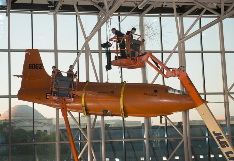 Using two lift machines, Museum employees work to secure the Bell X-1, an orange military monoplane, from the ceiling of the Boeing Milestones of Flight Hall onto the floor.