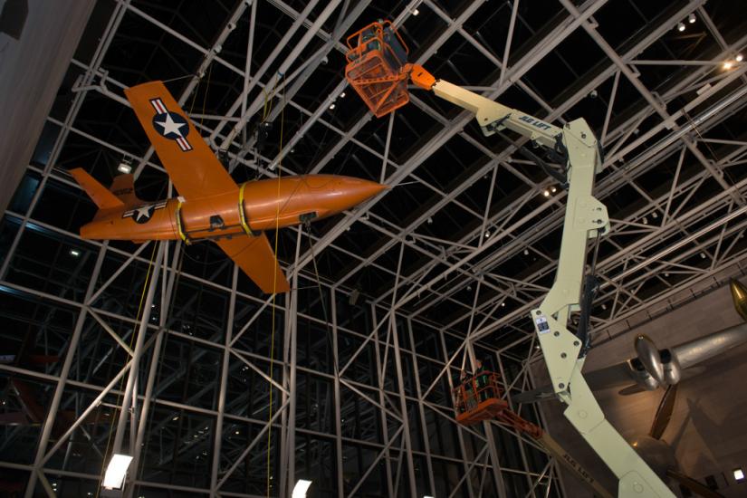 Using a singular lift machine, Museum employees work overnight to lower the Bell X-1, an orange military monoplane, from the ceiling to the floor of the Boeing Milestones of Flight Hall.